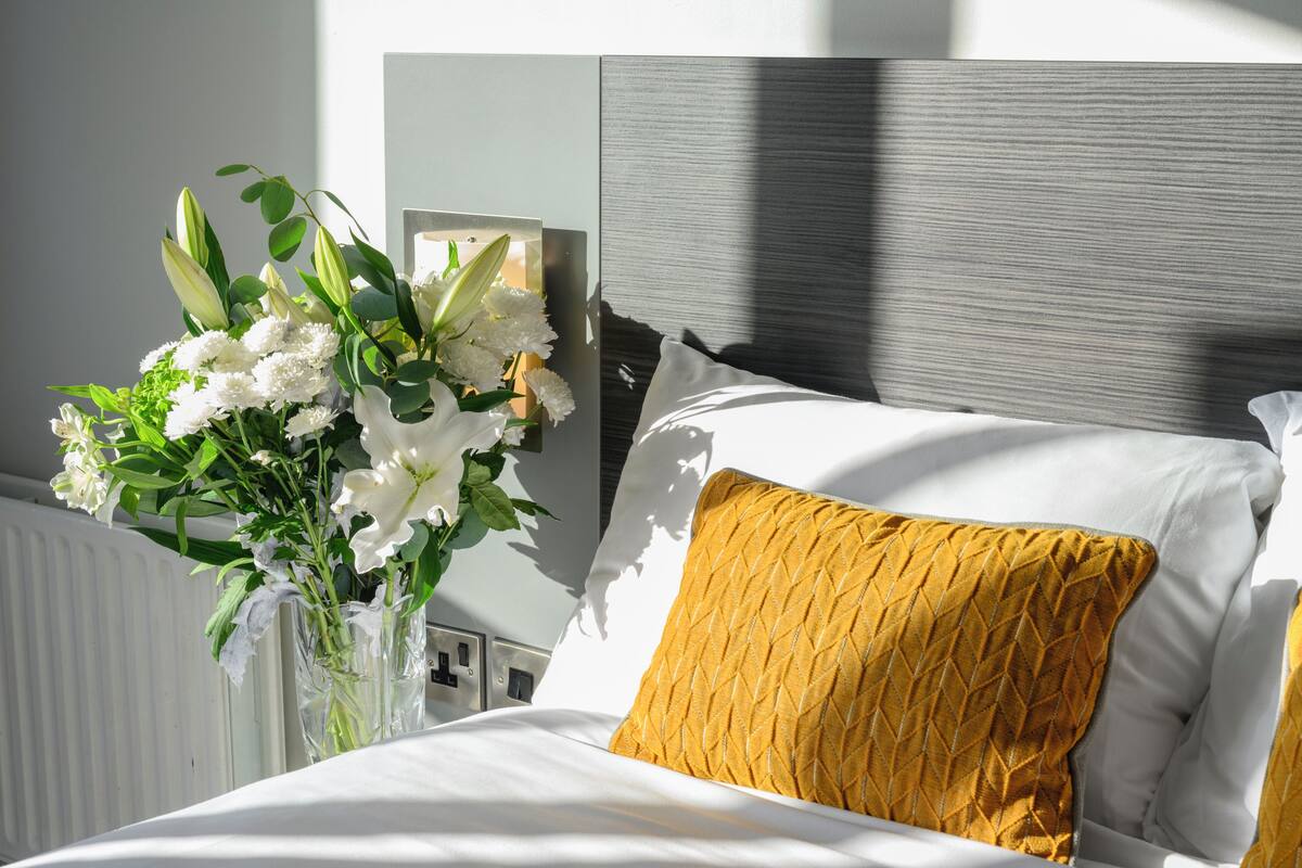 Ein ordentlich bezogenes Bett mit weißen Leinen und einem einzelnen senfgelben Kissen, eine Vase mit weißen Blumen und ein modernes Kopfteil.