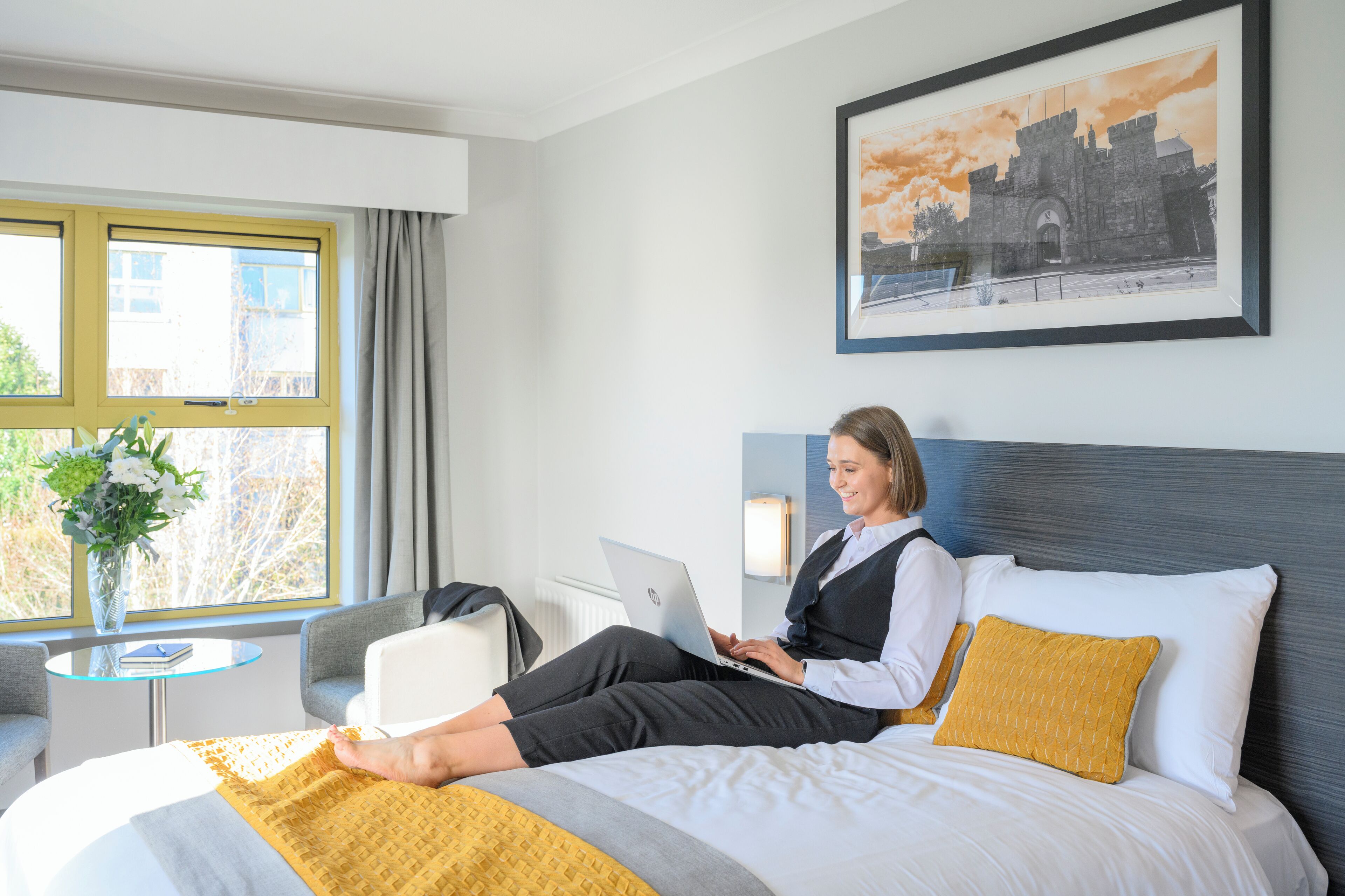 Une chambre d’hôtel avec un lit, un ordinateur portable, un vase de fleurs et une fenêtre avec des rideaux.