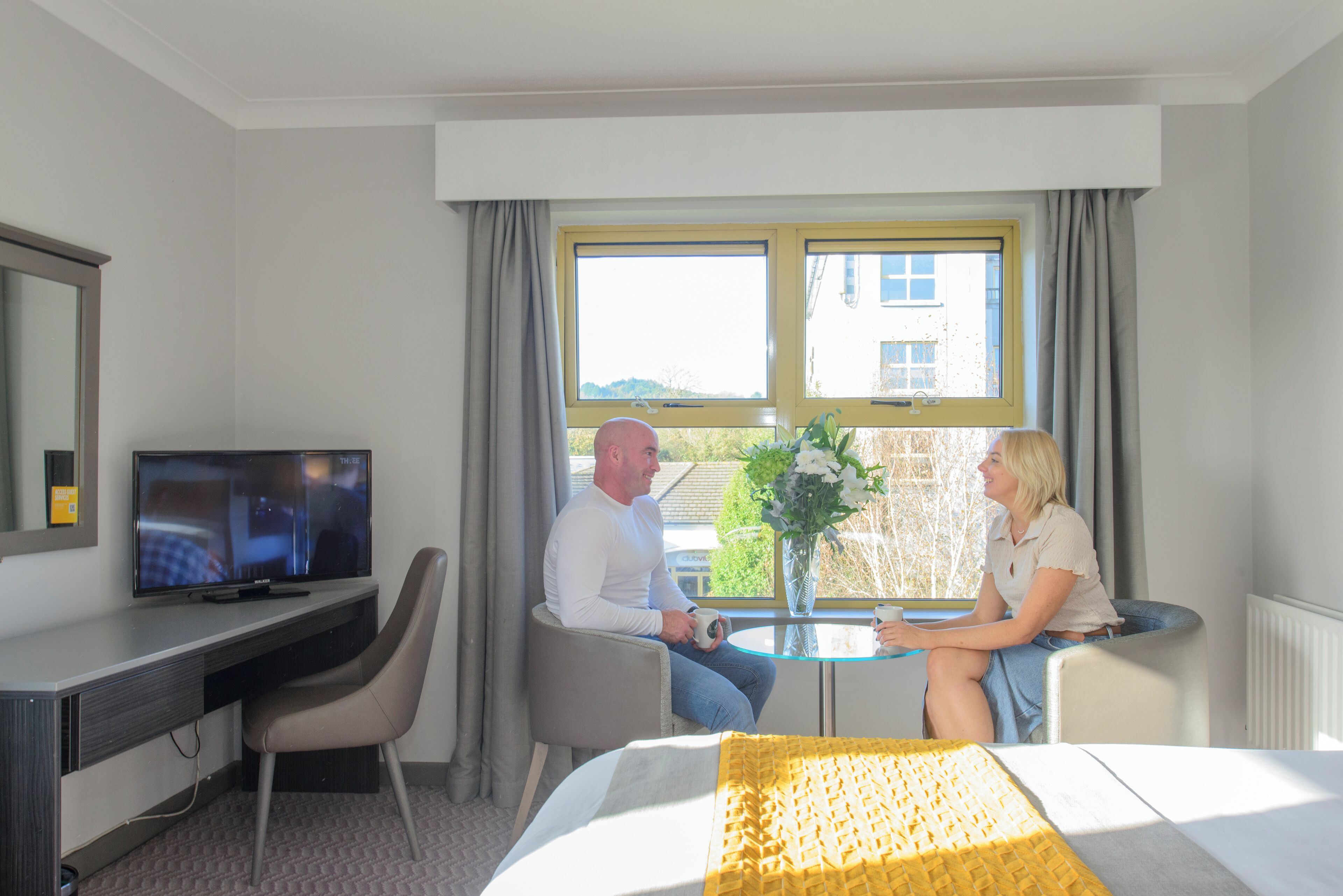 A hotel room with a bed, a desk with a TV, a window with curtains, and a small table with a vase of flowers.