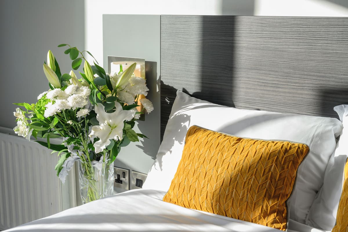 A bed with white bedding and a single yellow pillow, a vase with white flowers, and a wall-mounted light fixture.