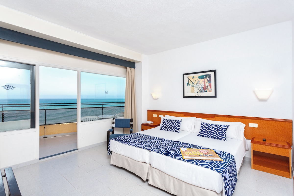 A hotel room with a large bed, a bedside table, a nightstand, a chair, a window with a view of the sea, and a framed picture on the wall.