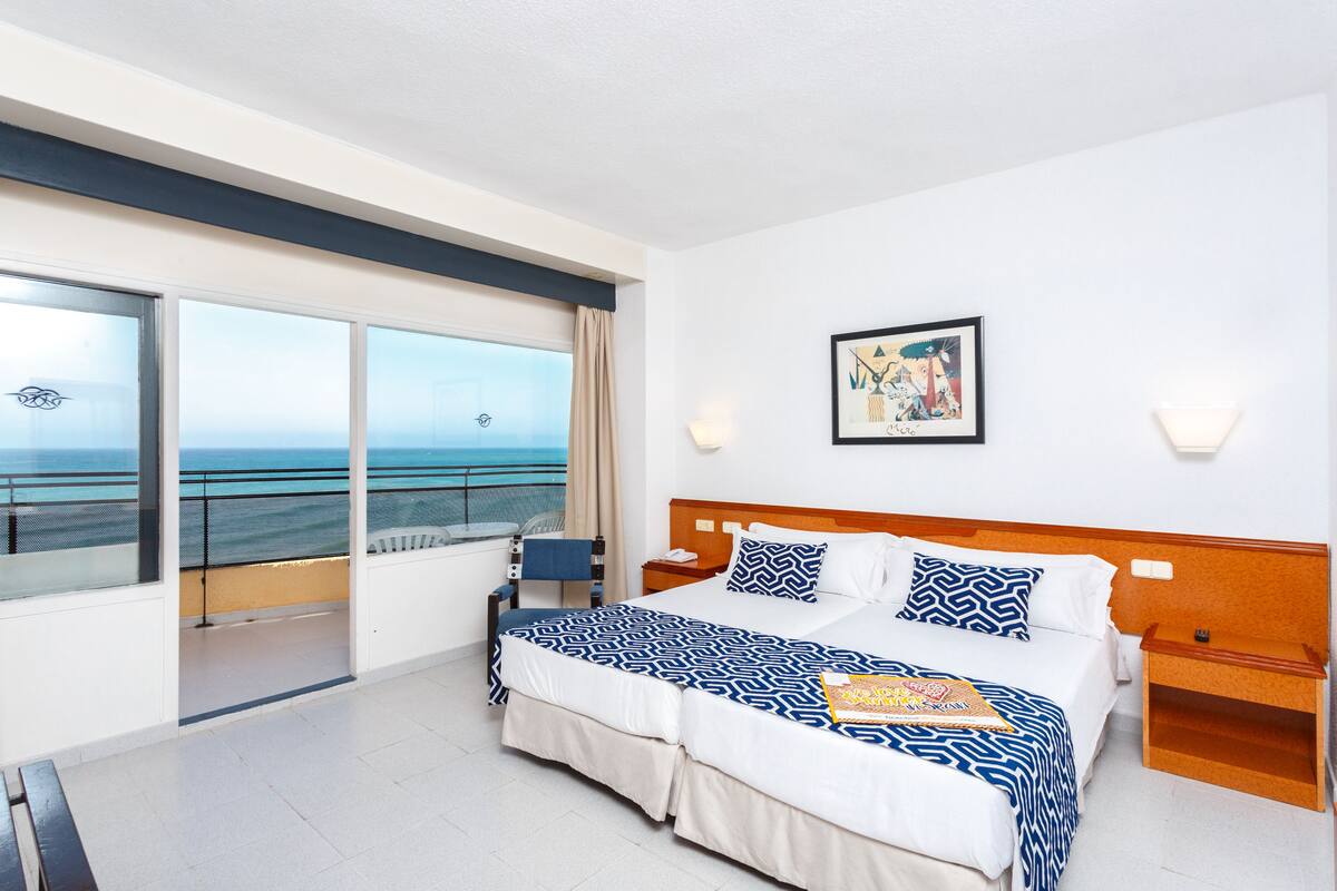 Quarto de hotel com uma cama grande, uma mesinha de cabeceira, uma cadeira e uma janela com vista para o mar, além de um quadro na parede.