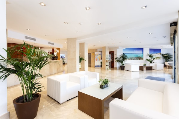 A modern hotel lobby with white sofas, a large potted plant, and a view of the ocean through the windows. - Globales Gardenia (Fuengirola)