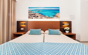 A double bed with a patterned bedspread, two pillows, a headboard, bedside tables with lamps, and a wall-mounted painting of a coastal landscape. - Globales Aparthotel Bouganvilla (Sant Llorenc des Cardassar)