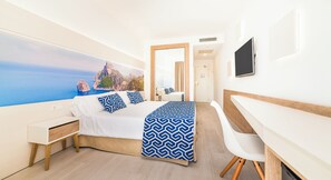 Habitació d'hotel amb un llit, una escriptori, una cadira, una televisió i una finestra gran amb vista al mar.