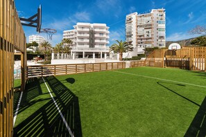 Una cancha deportiva con aro de baloncesto, rodeada de vallas de madera, y edificios al fondo.