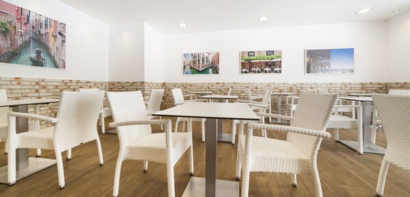 A modern dining area with white wicker chairs and tables, brick accents on the walls, and cityscape artwork.