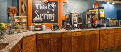 A kitchen area with a chalkboard sign that reads "Think Spring," a waffle maker, a coffee machine, and a display case with pastries.