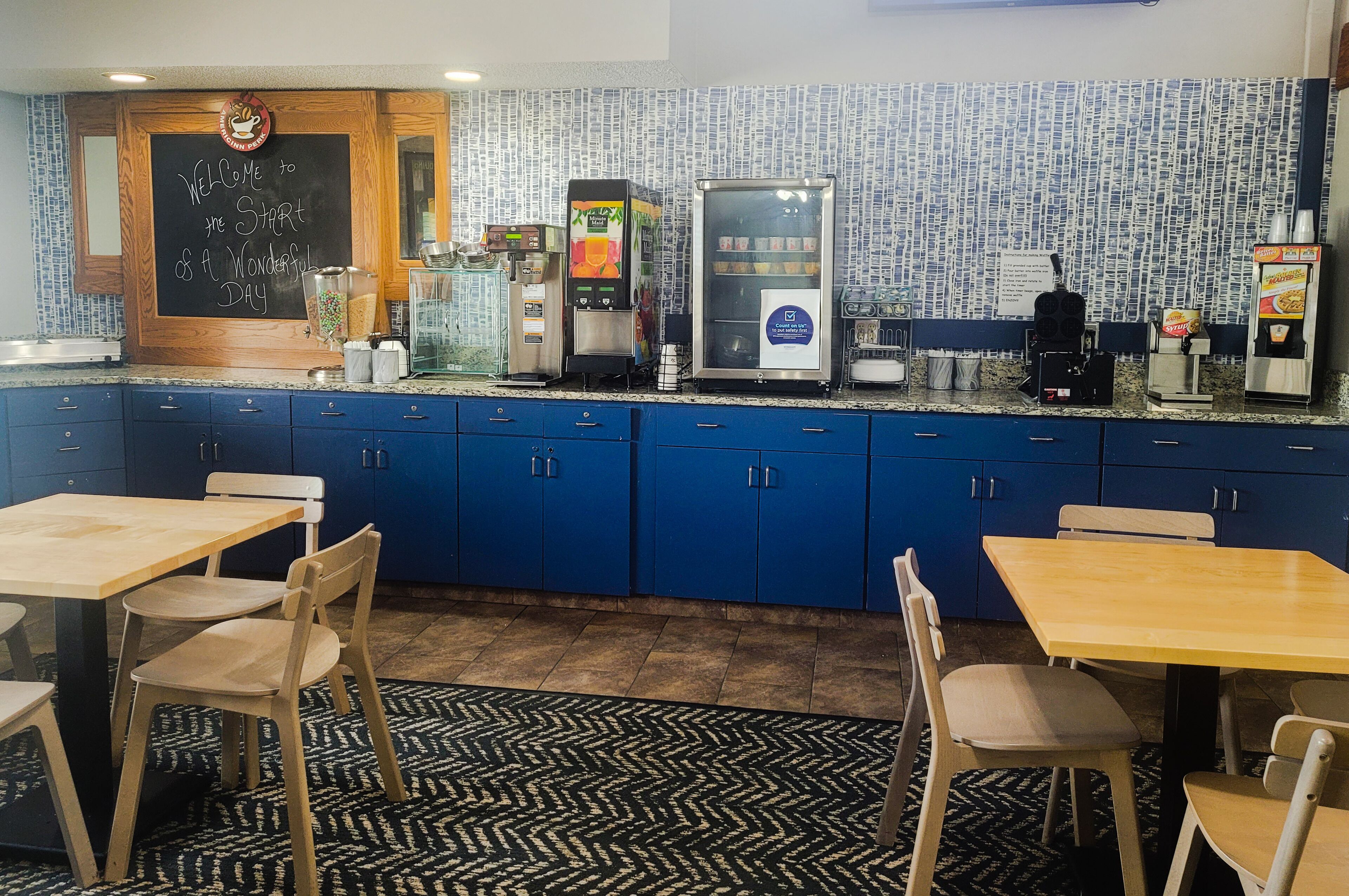 Un restaurante con armarios azules, una pizarra con un mensaje de bienvenida, una máquina de café, un refrigerador exhibidor y un dispensador de café.