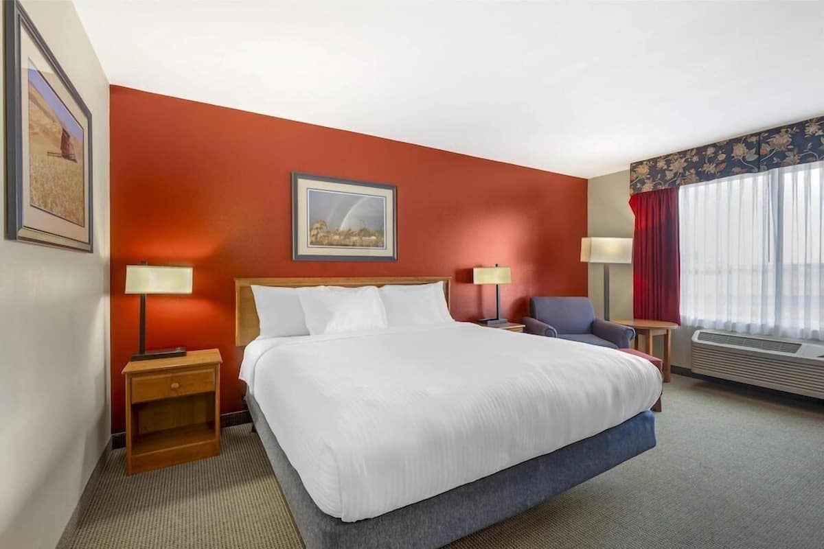 Ein Hotelzimmer mit einem großen Bett, zwei Nachttischen, einem Sessel, einem Fenster mit Vorhängen und einem Bild an der Wand.