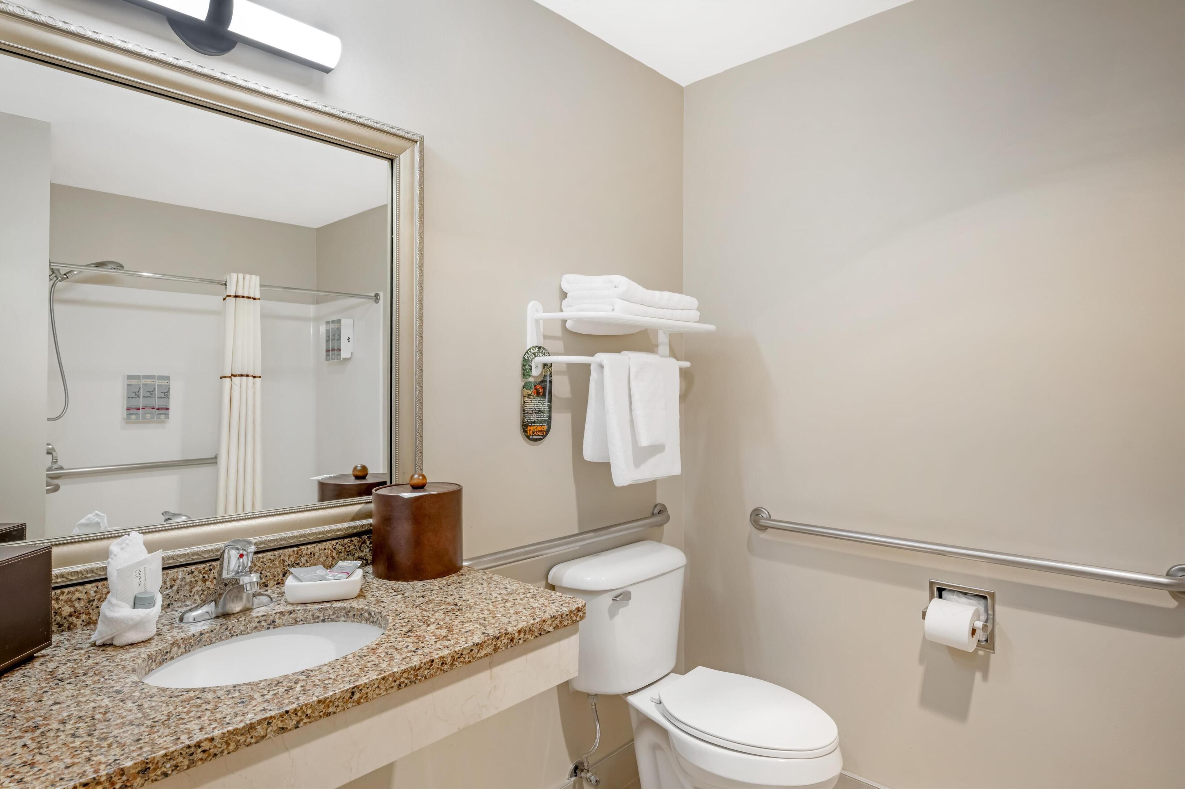 Une salle de bain dotée d’un lavabo avec un plan de travail en granit, de toilettes blanches, d’un porte-serviettes avec des serviettes pliées, d’un sèche-mains et d’un miroir avec un distributeur de serviettes.