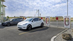 Dois carros elétricos Tesla estacionados numa estação de carregamento, com um céu claro ao fundo.