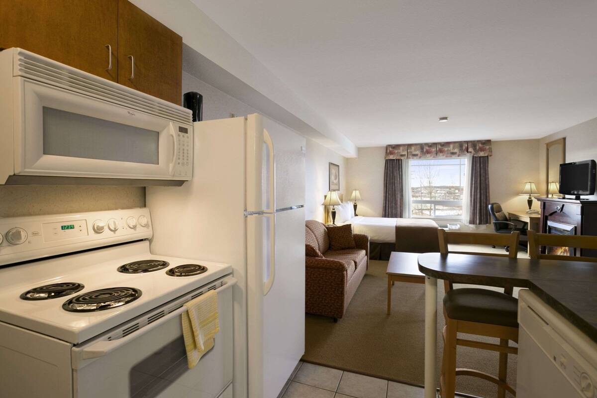 A compact kitchen with a stove, microwave, and refrigerator. There is a dining area with a table and chairs, and a sofa in the background.