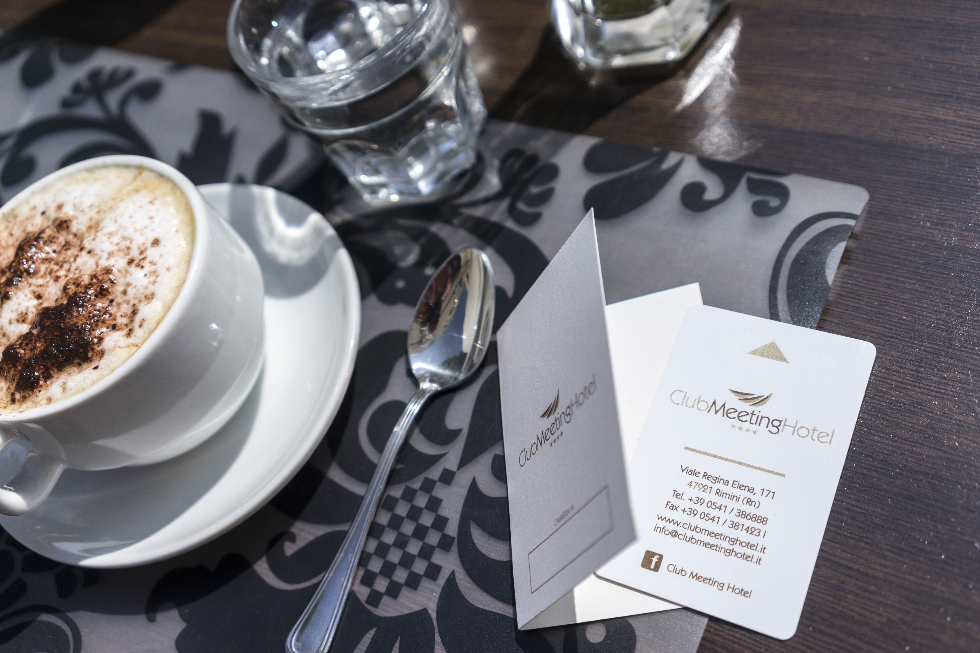 Eine Visitenkarte des ClubMeeting Hotels mit Kontaktdaten, einem Löffel, einem Glas Wasser und einer Tasse Kaffee auf einem Tisch.