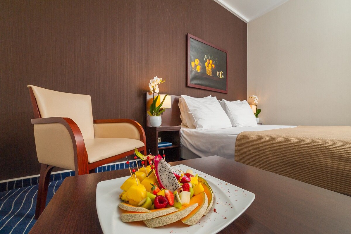 A hotel room with a bed, a chair, a table with fruit, and a painting on the wall.