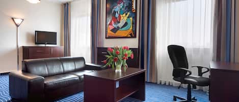 A hotel room with a leather sofa, a small wooden coffee table with a vase of red flowers, a black office chair, a television on a wooden cabinet, and a large abstract painting on the wall.