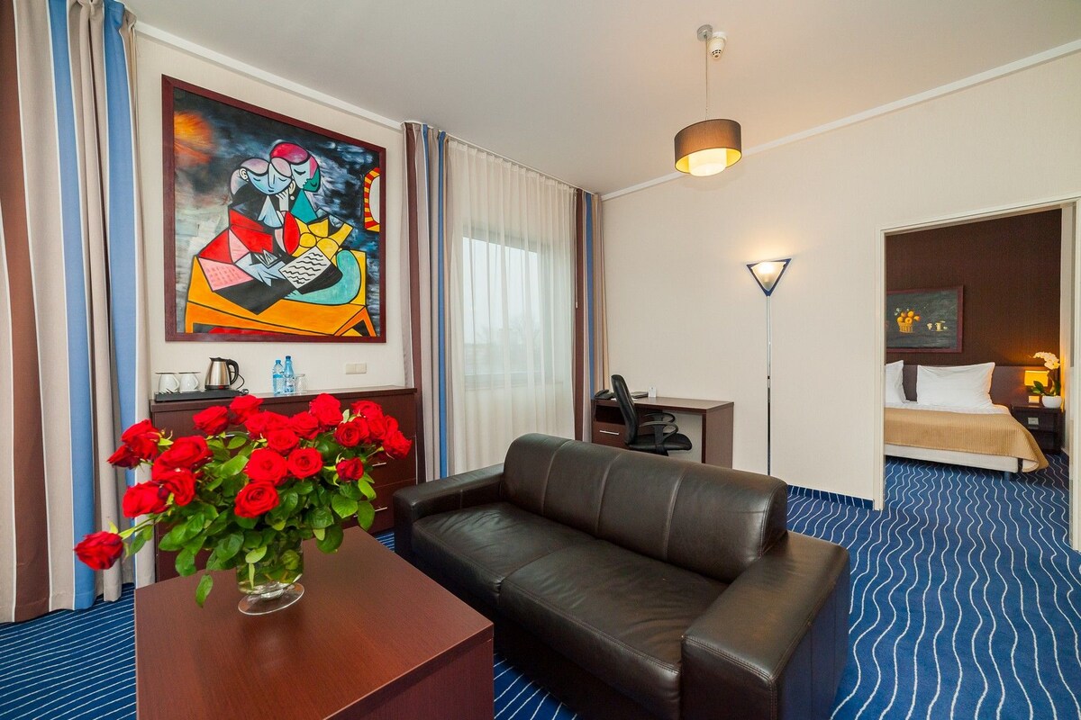 A hotel room with a sofa, a desk with a vase of red roses, a painting on the wall, a bed with a blue patterned bedspread, and a nightstand with a lamp.