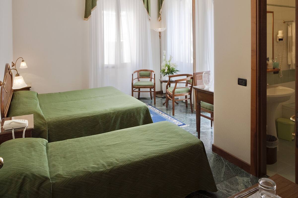 Habitación de hotel con dos camas, un escritorio, una silla, una lámpara y un baño visible a través de una puerta abierta.