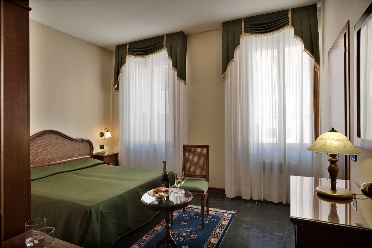 Habitación de hotel con cama, una mesa con botella y vasos, una silla y un escritorio con lámpara.