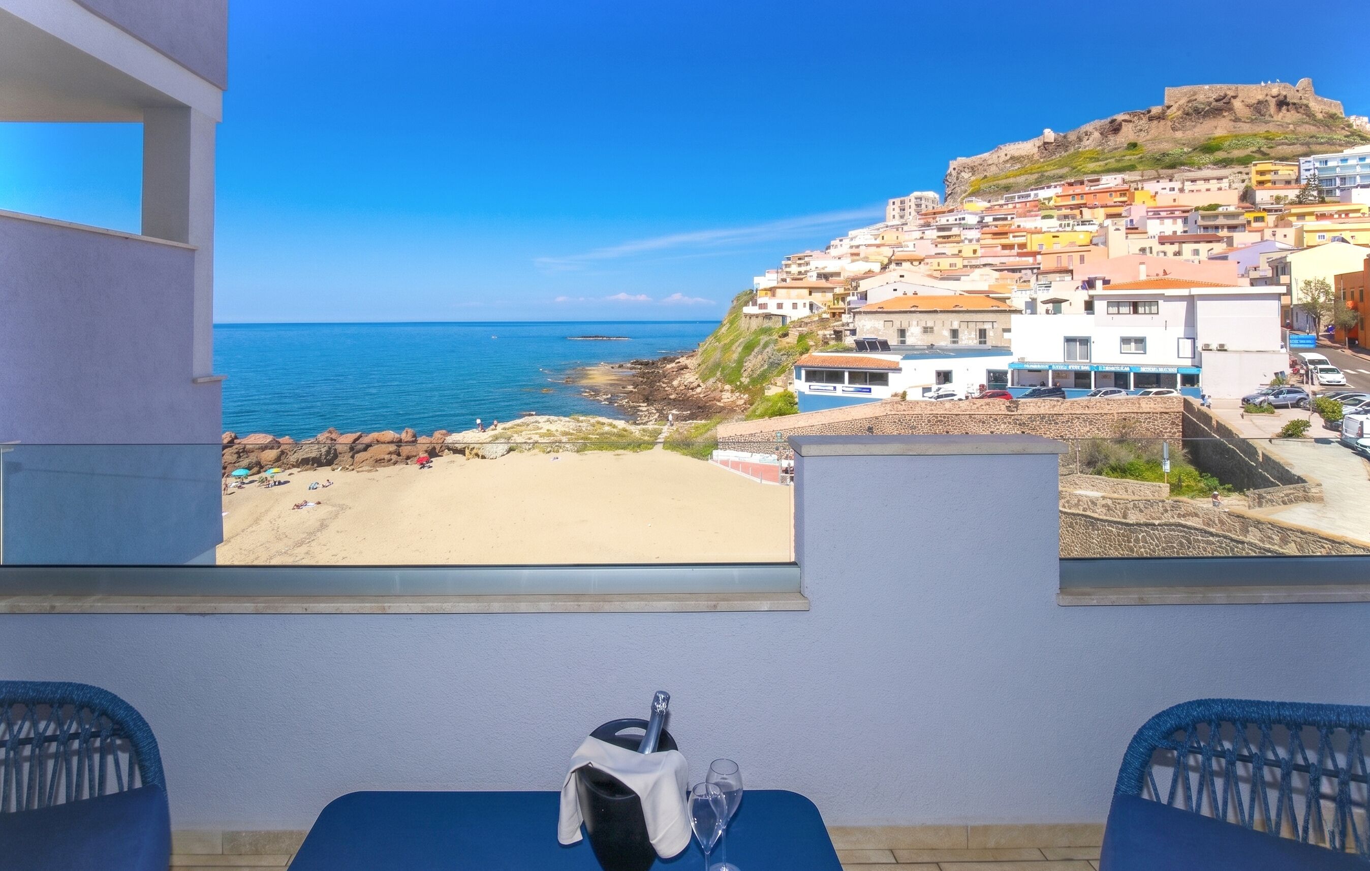 Un balcón con vista a un pueblo costero, una mesa con una silla y una ventana que da al mar y edificios.