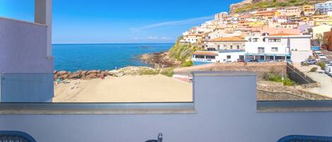 Un balcón con vista a un pueblo costero, una mesa con una silla y una ventana que da al mar y edificios.