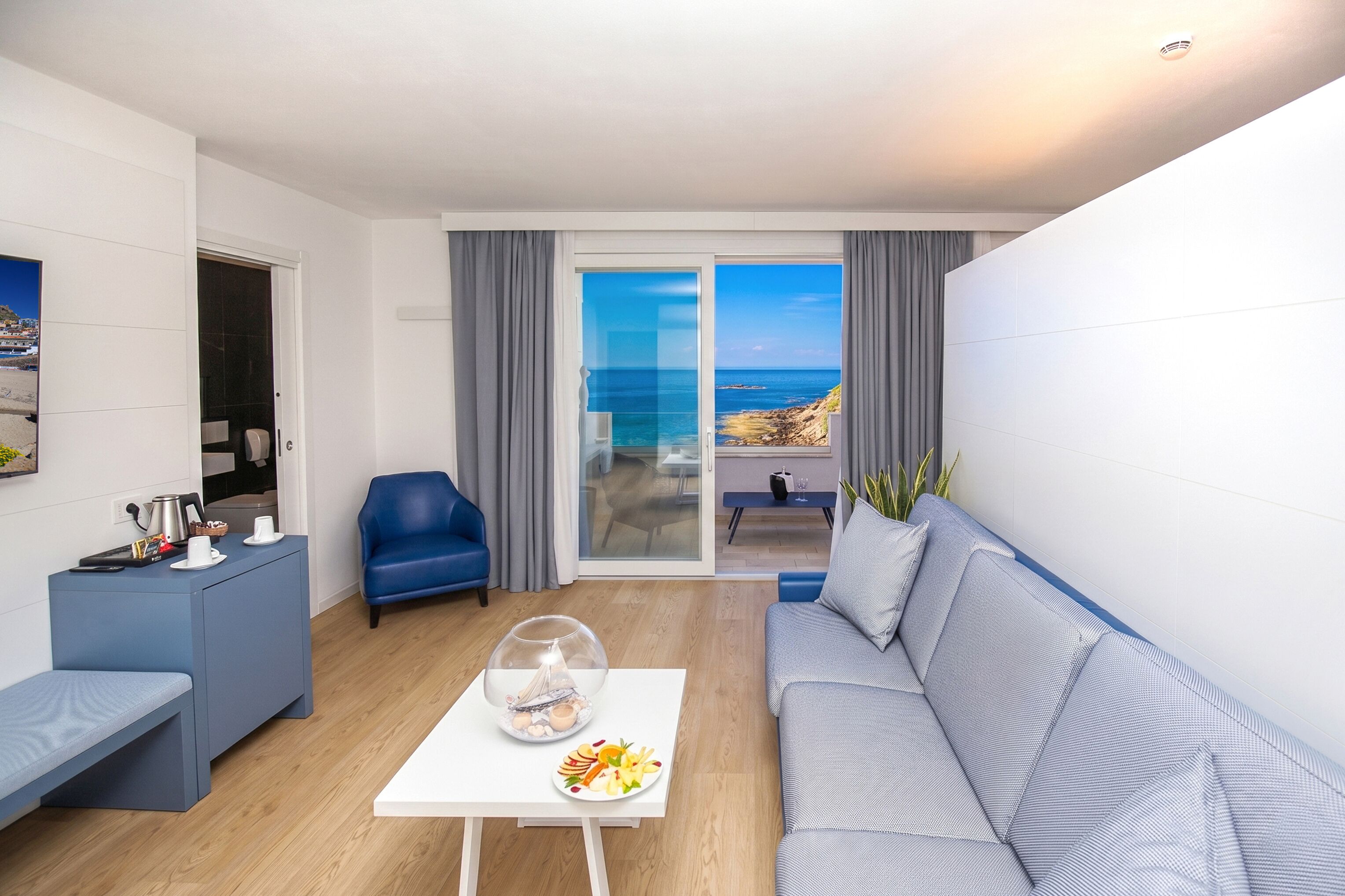 Una sala de estar moderna con un sofá, una mesa de centro con un objeto decorativo, un sillón azul y vista al océano a través de un ventanal.