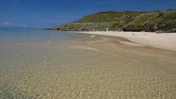Una playa con aguas cristalinas poco profundas, una orilla arenosa y una colina cubierta de hierba al fondo.