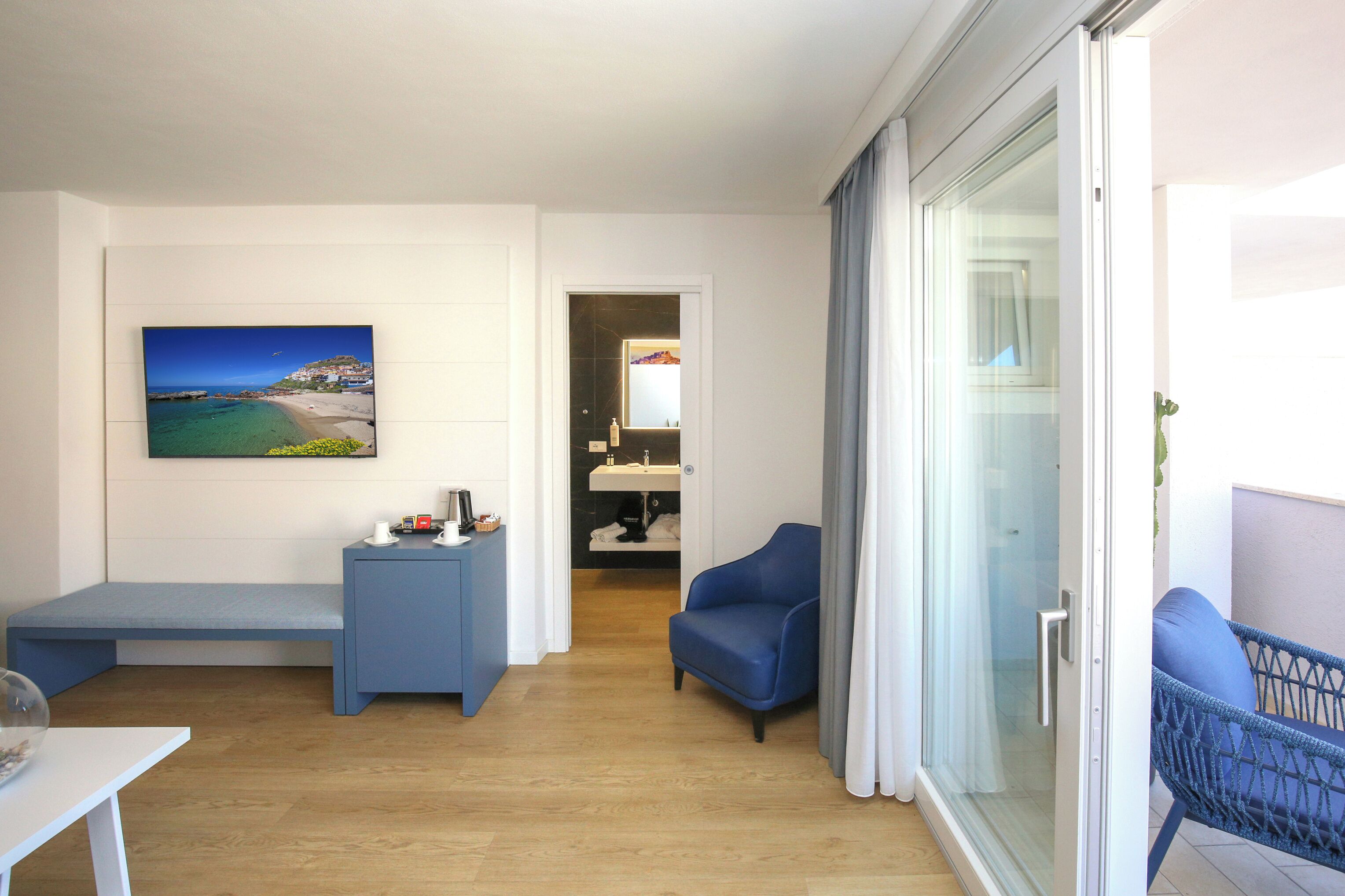 Una sala de estar moderna con piso de madera, un sofá azul, un banco, una puerta de vidrio que da a un balcón y un cuadro con una escena de playa en la pared.