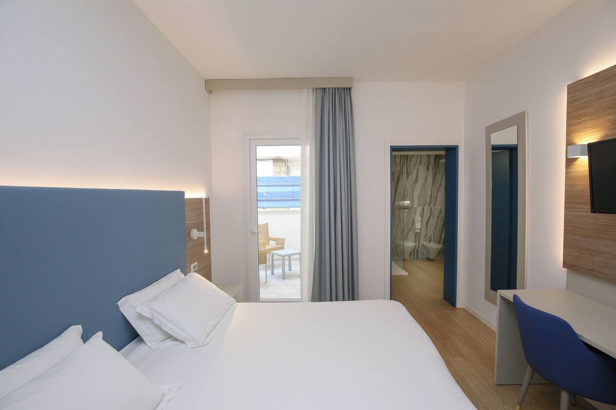 A hotel room with a bed, a desk, a chair, a TV, and a view of the outside.