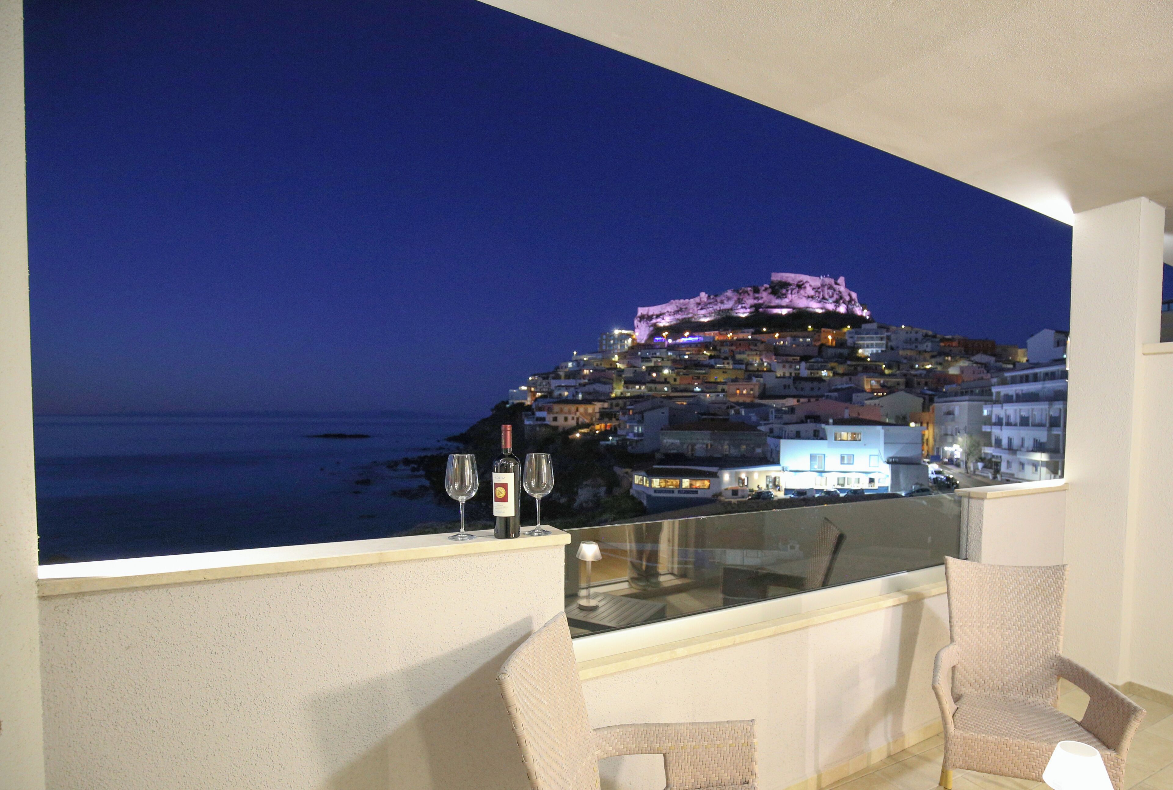 A balcony with a view of a coastal town at night, featuring a chair and two wine glasses.