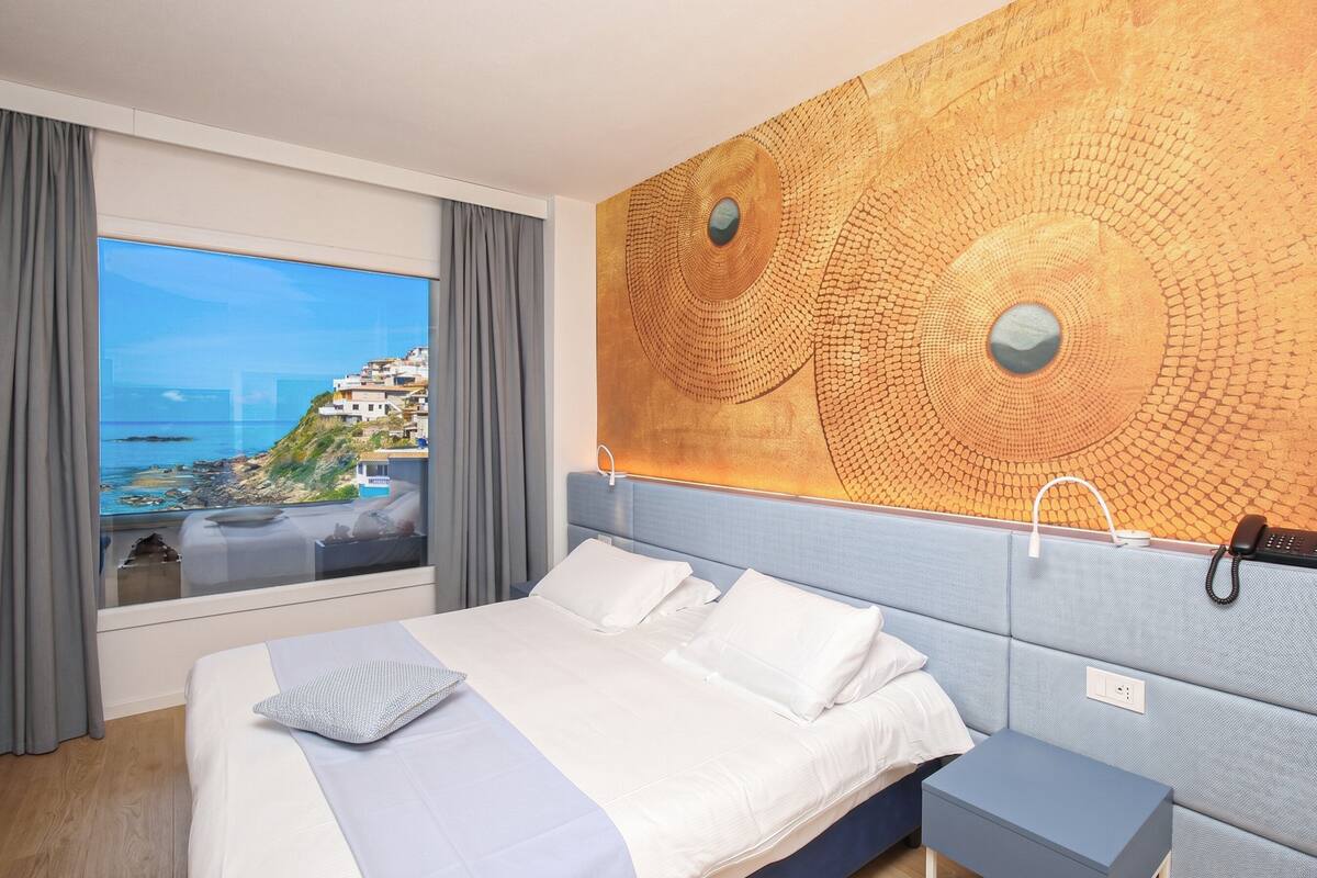 A hotel room with a bed, a nightstand, a large window with a view of a coastal landscape, and a wall with a textured artwork.