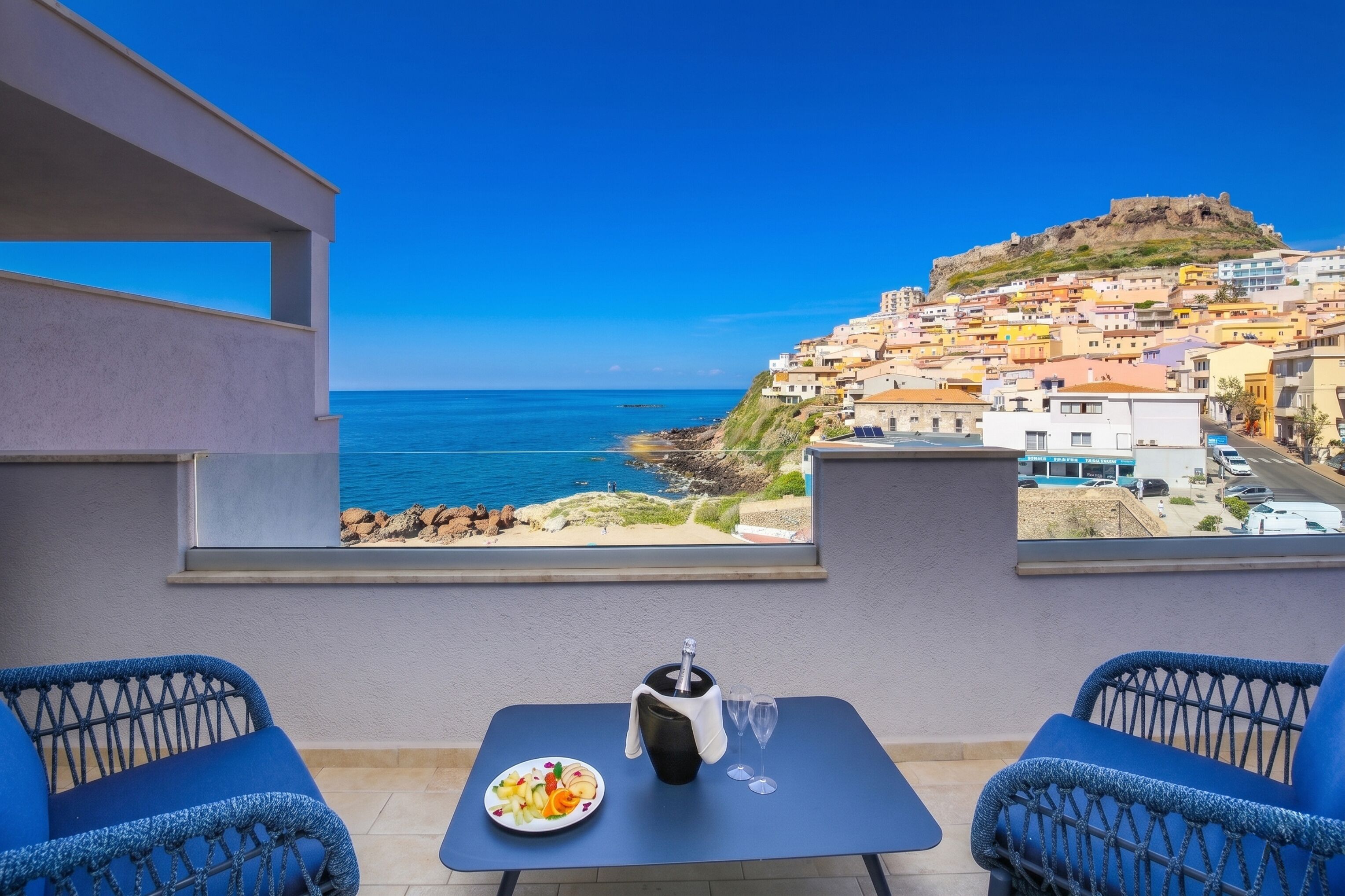 Un balcón con una mesa, dos sillas, una botella y un plato de comida, con vistas a un pueblo costero con edificios y una colina.