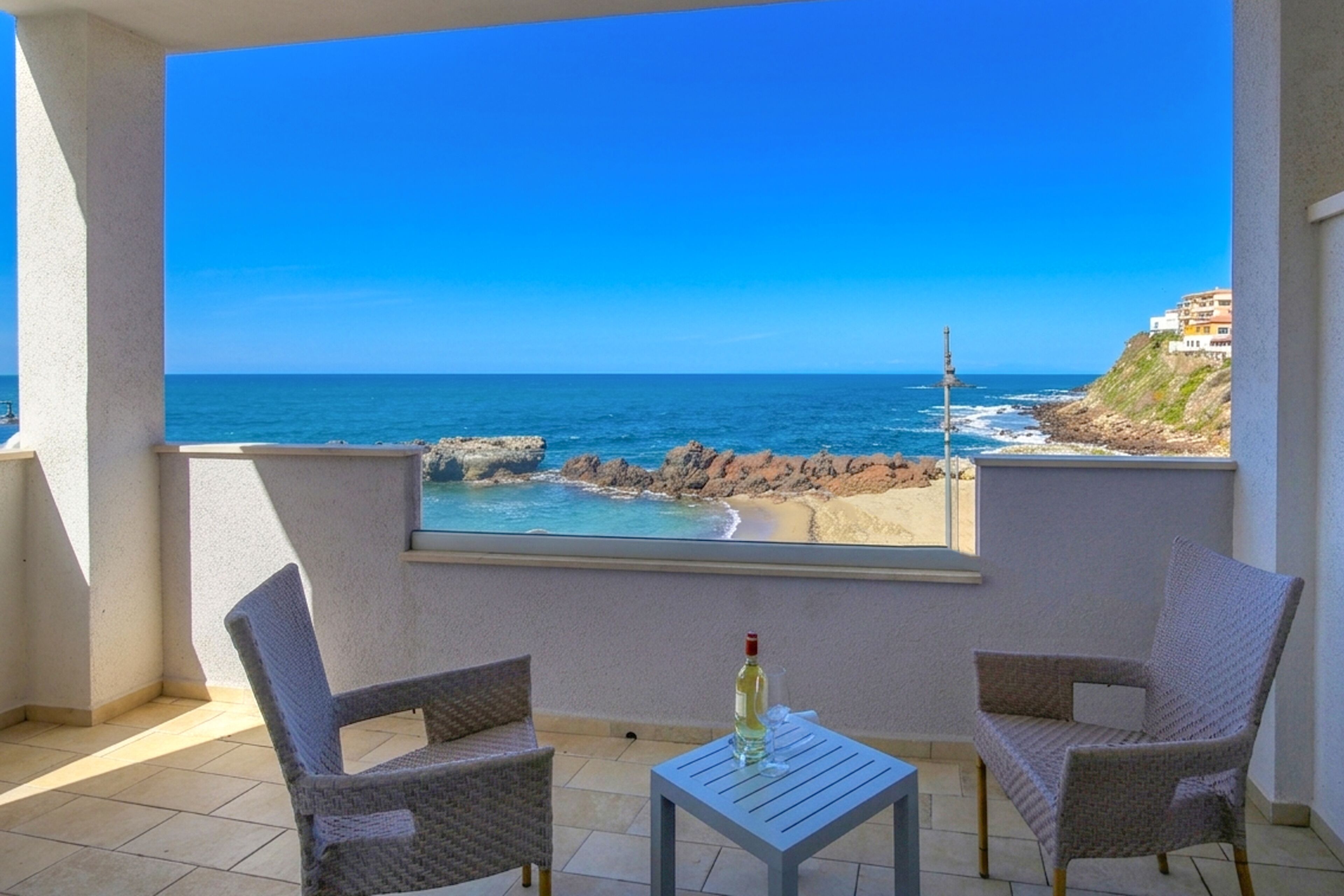 Un balcon avec deux fauteuils en osier, une petite table et une bouteille. La vue comprend une plage et une côte rocheuse.