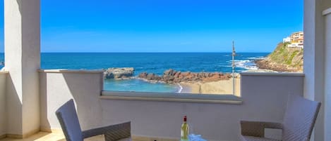 Un balcon avec deux fauteuils en osier, une petite table et une bouteille. La vue comprend une plage et une côte rocheuse.