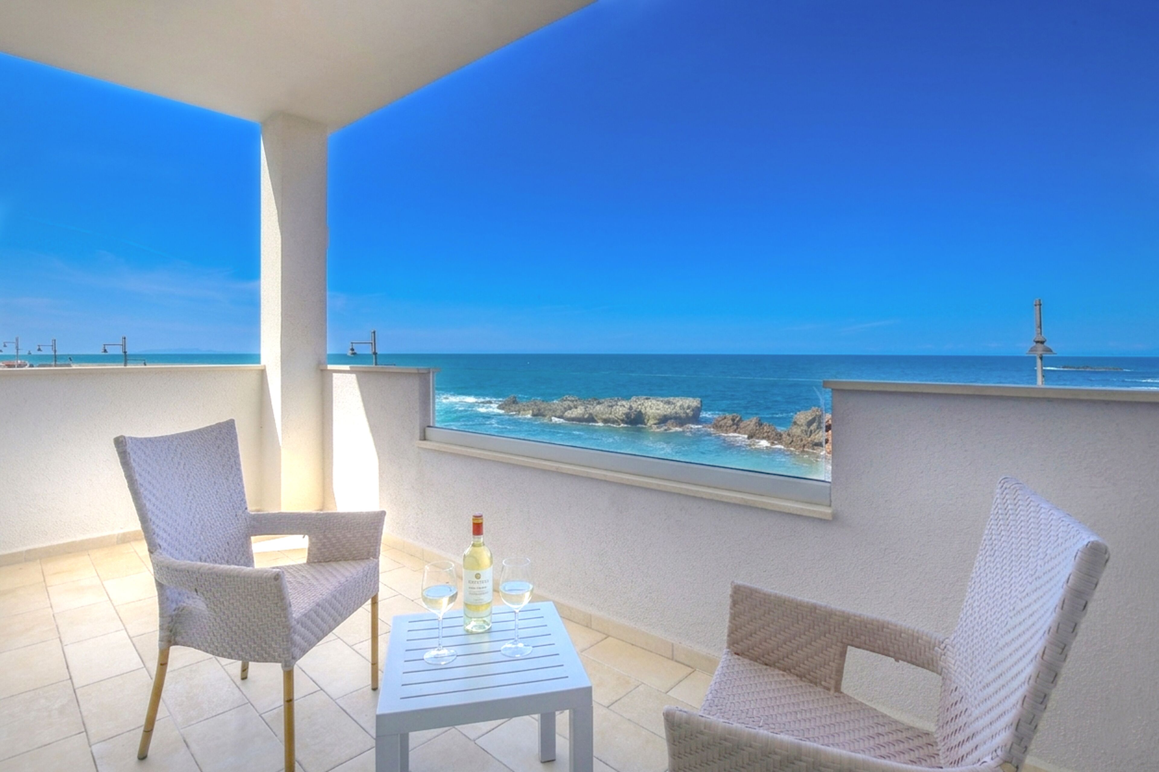 A balcony with two wicker chairs, a small table, and a bottle of wine. The view includes the sea and rocky coastline.