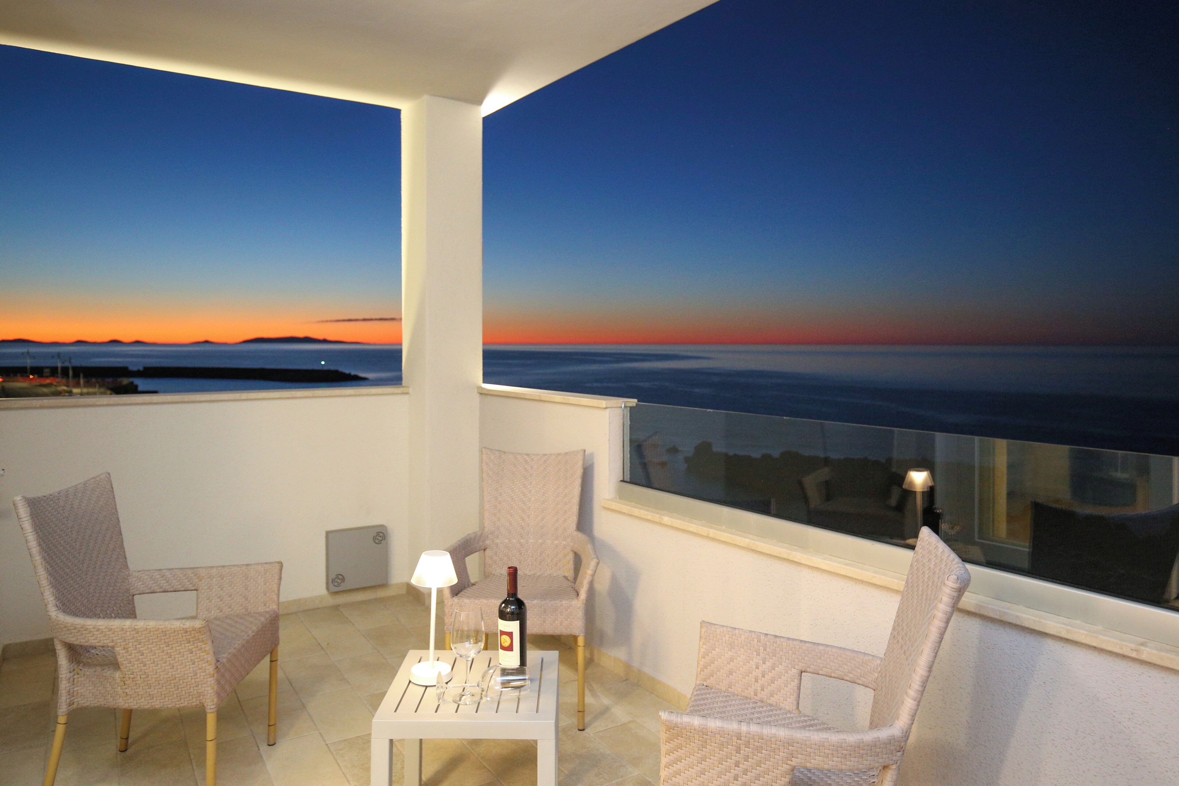 Un balcon avec deux fauteuils en osier, une petite table sur laquelle se trouvent une bouteille de vin et des verres, et une vue sur le coucher de soleil sur l’eau.