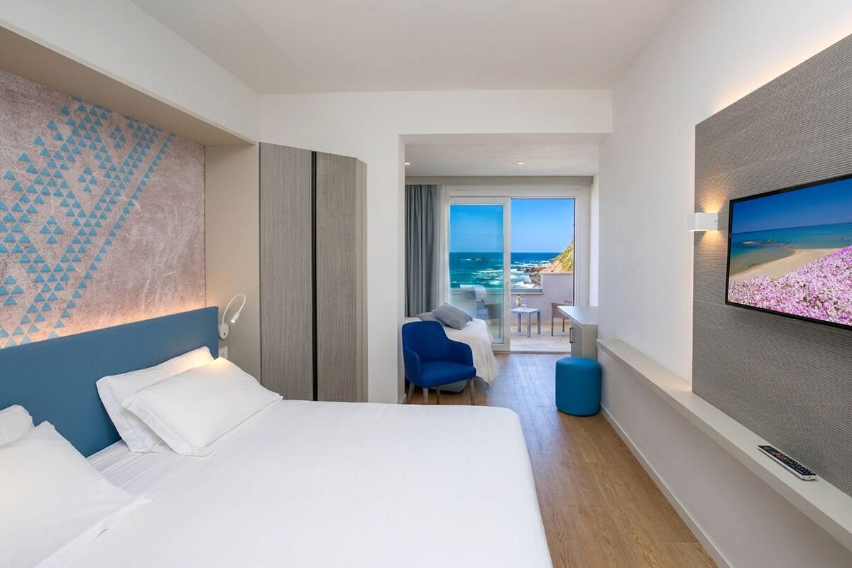 A hotel room with a bed, a TV, a view of the ocean, and a balcony.
