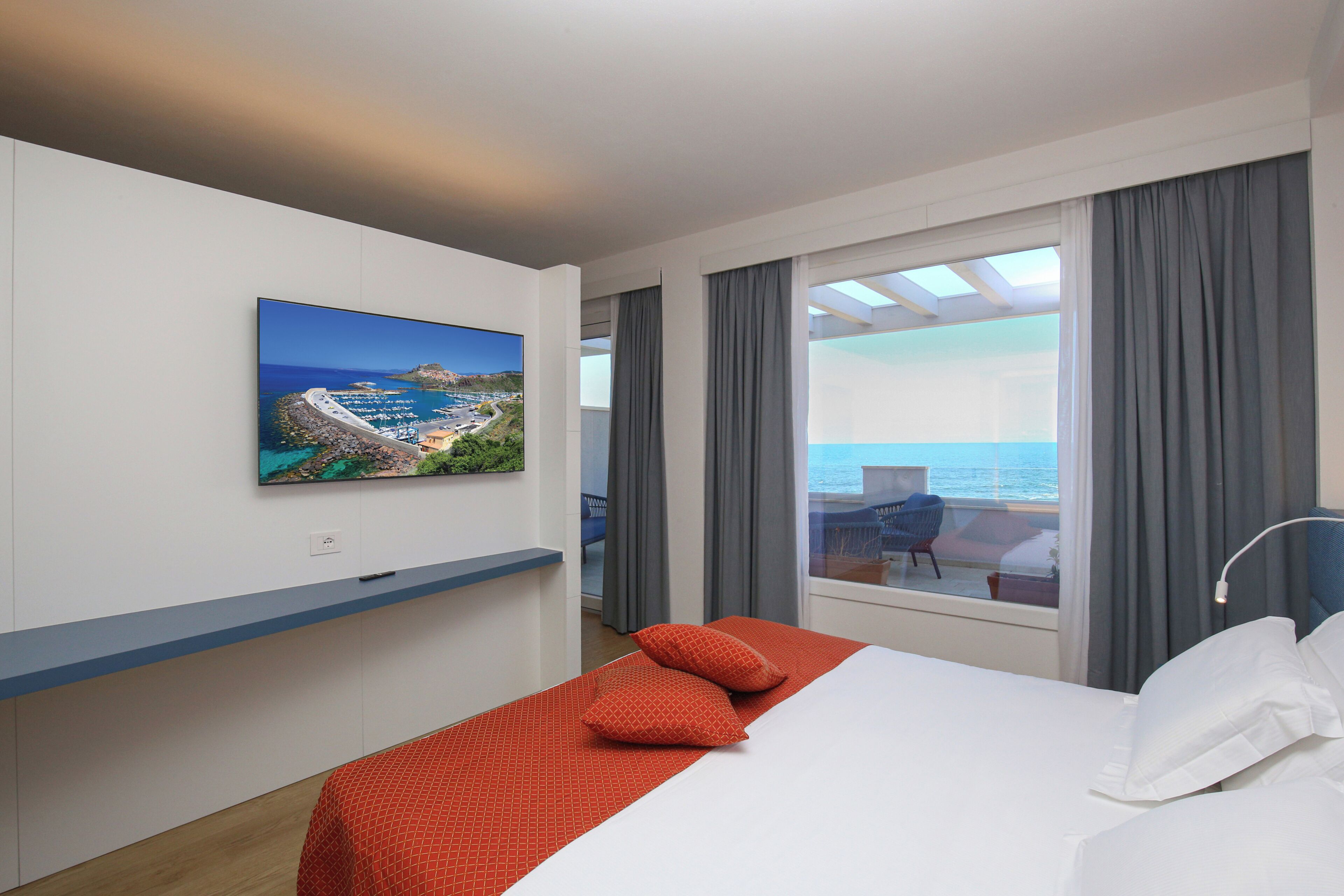A hotel room with a bed, a TV mounted on the wall, a balcony with a view of the sea, and a window with curtains.