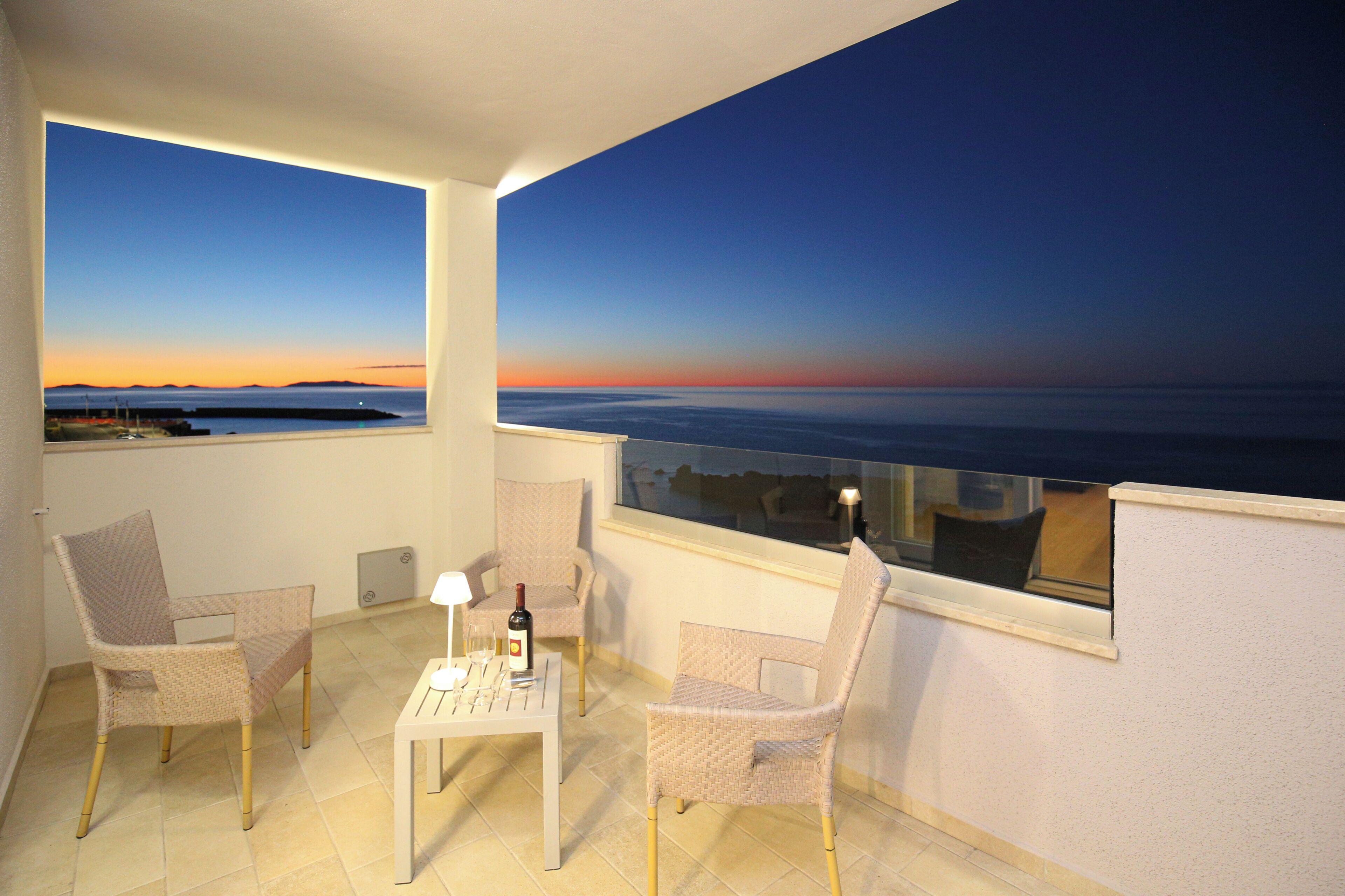 A balcony with two wicker chairs, a small table with a bottle and glasses, and a view of the sea at sunset.