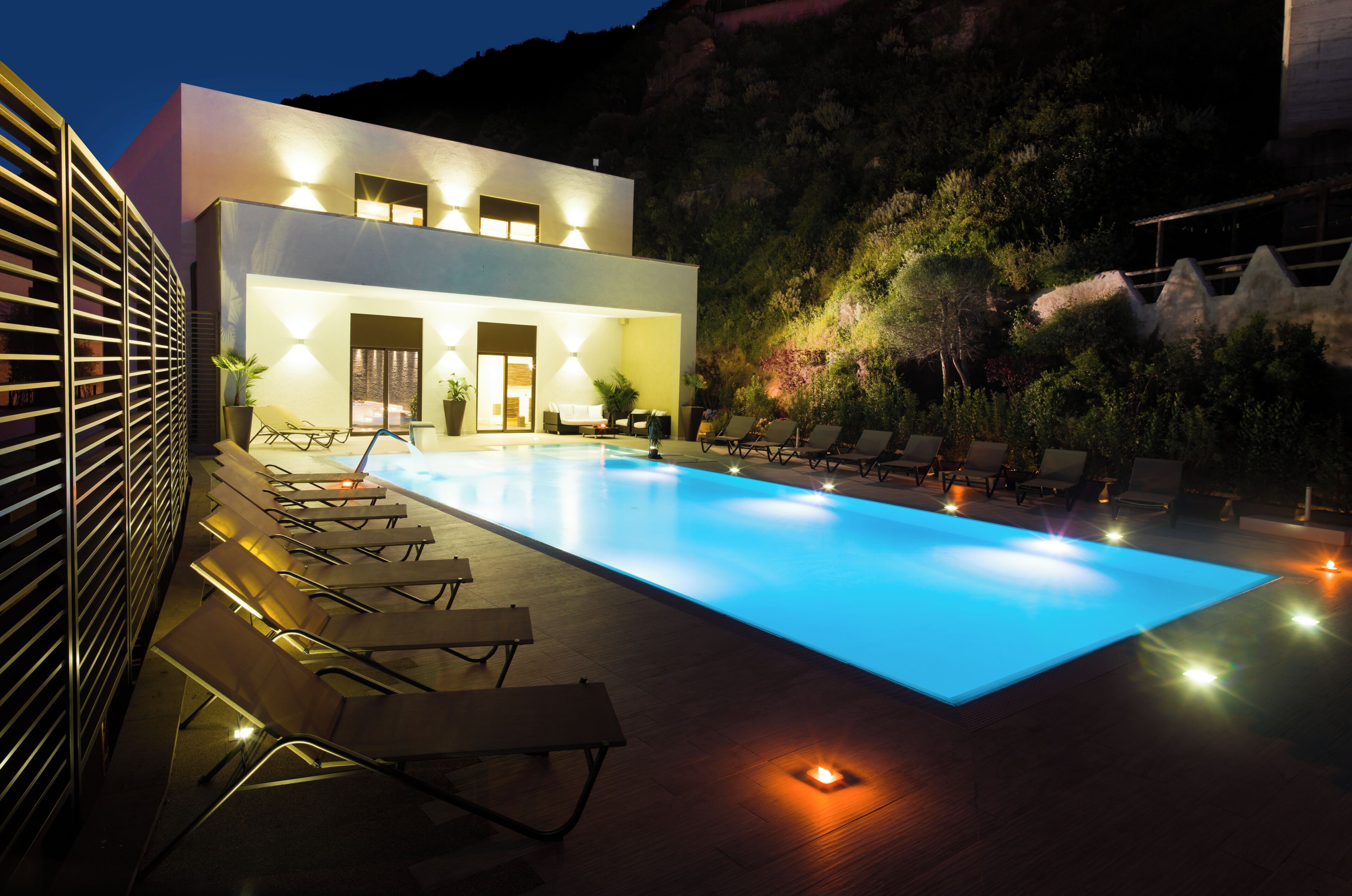 A modern pool area with lounge chairs, a pool, and a building with exterior lighting.