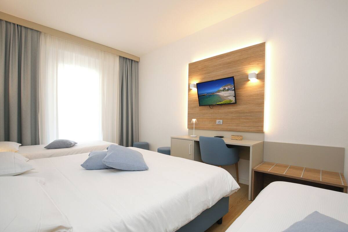 A hotel room with two beds, a desk, a chair, a TV mounted on the wall, and a window with curtains.