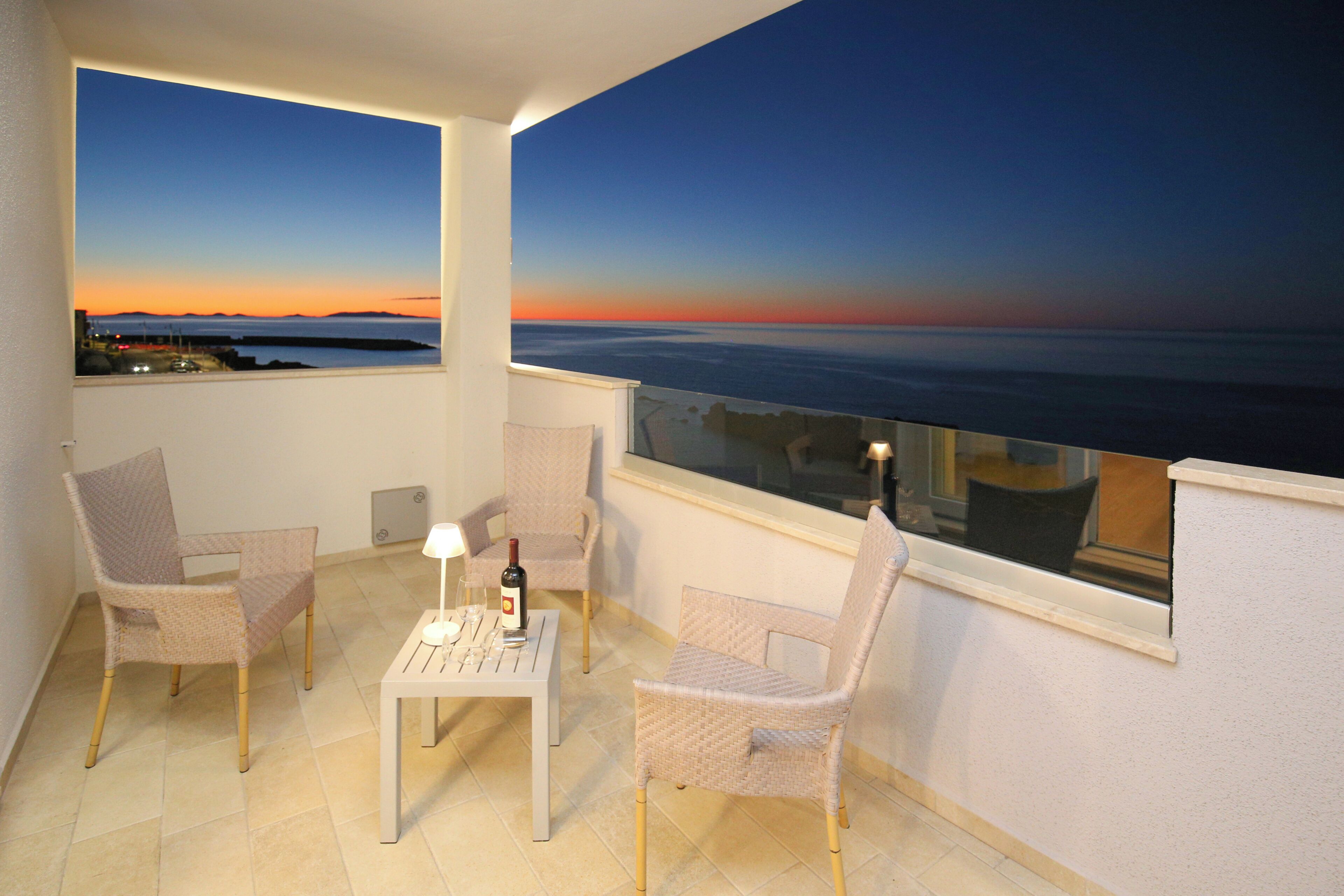 A balcony with two wicker chairs, a small table with a bottle and glass, and a view of the sunset over the water.