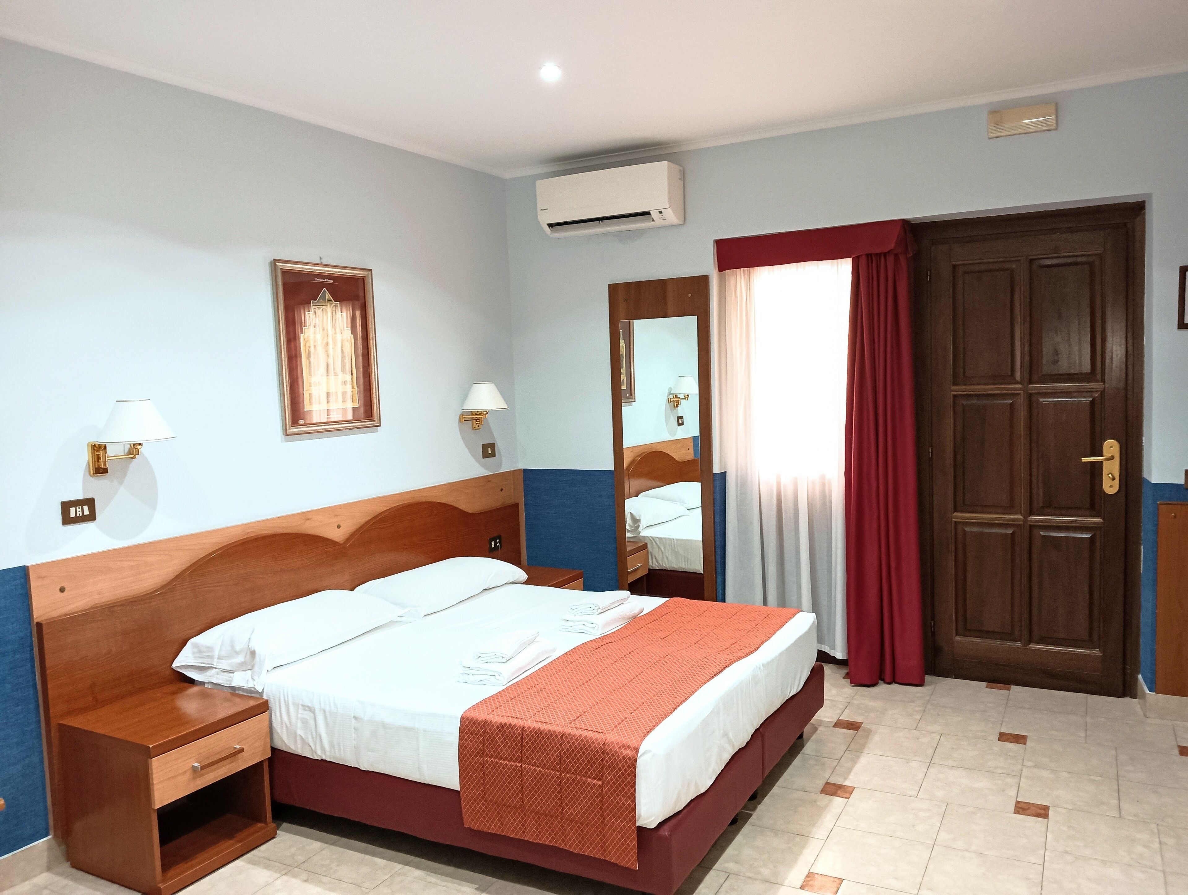 Une chambre d’hôtel avec un lit en bois, une table de chevet, un miroir, un appareil de climatisation et une porte.