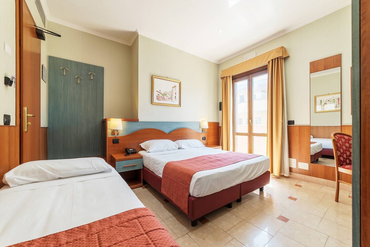 Een hotelkamer met twee bedden, een houten garderobe, een nachtkastje, een spiegel en een deur met gordijn.