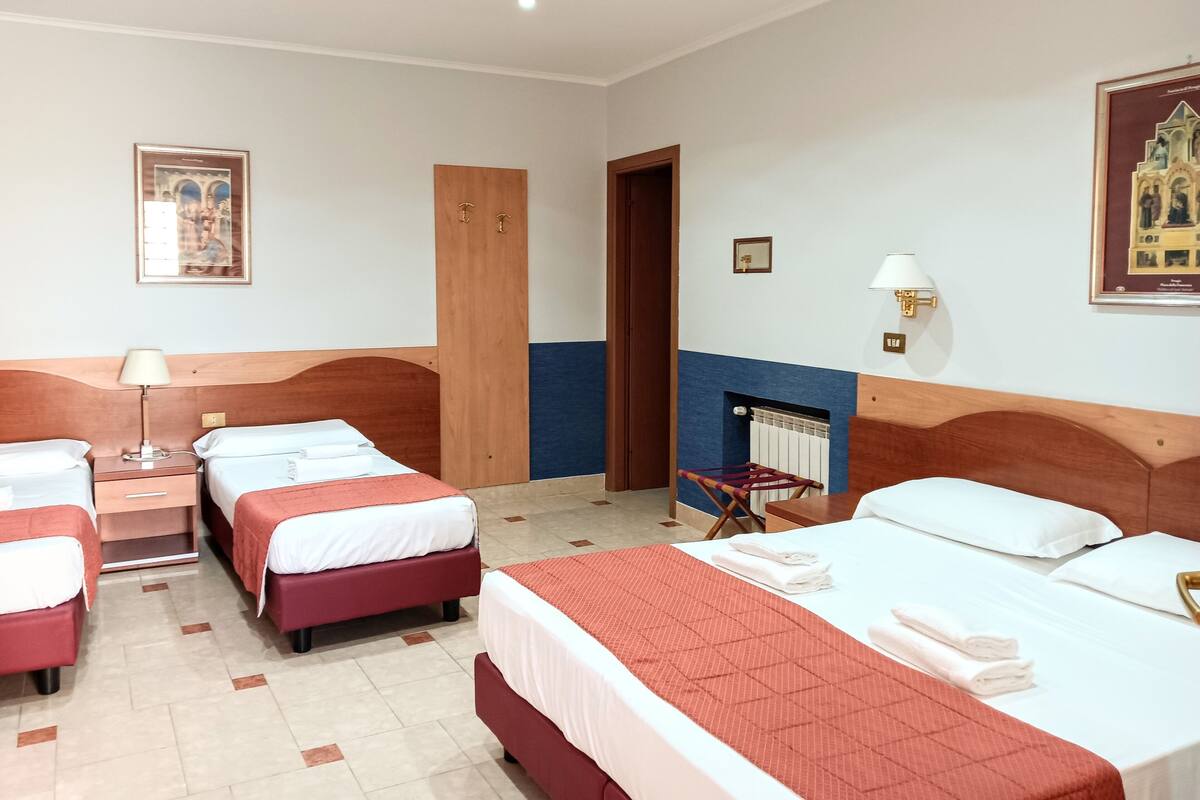 Een hotelkamer met vier bedden, een houten hoofdbord, een klein tafeltje met een lamp, een ingelijst schilderij aan de muur en een open haard.