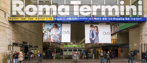 A busy train station with "RomaTermini" signage, multiple advertisements, and a clear view of the platforms.