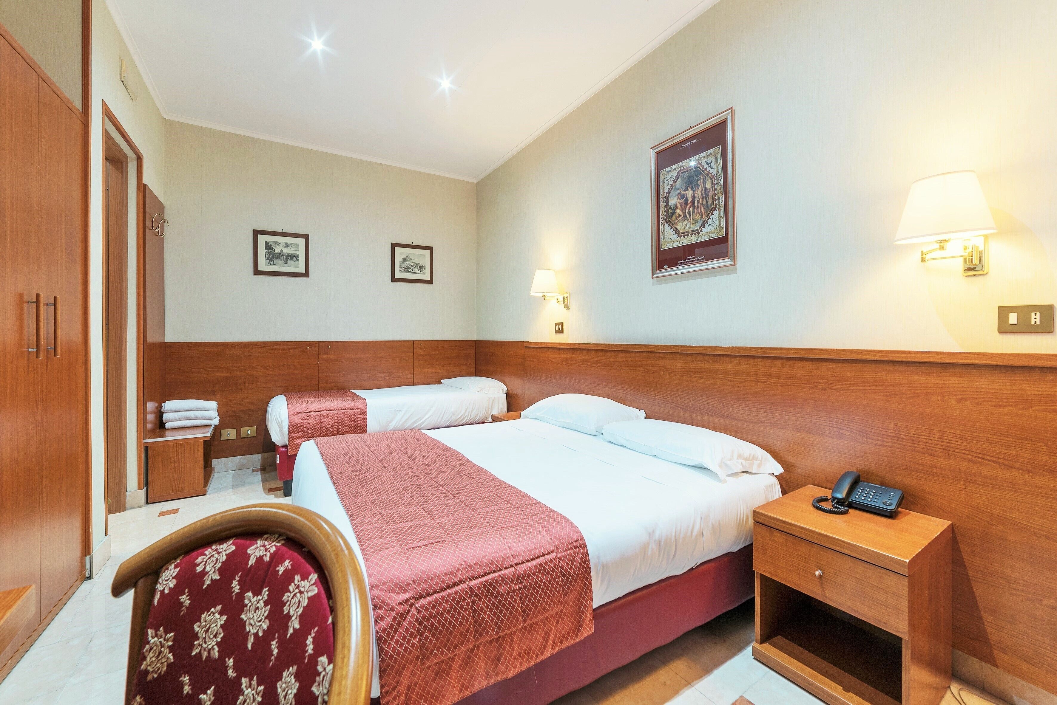 A hotel room with two beds, a wooden nightstand with a phone, a chair, and a wall-mounted lamp.