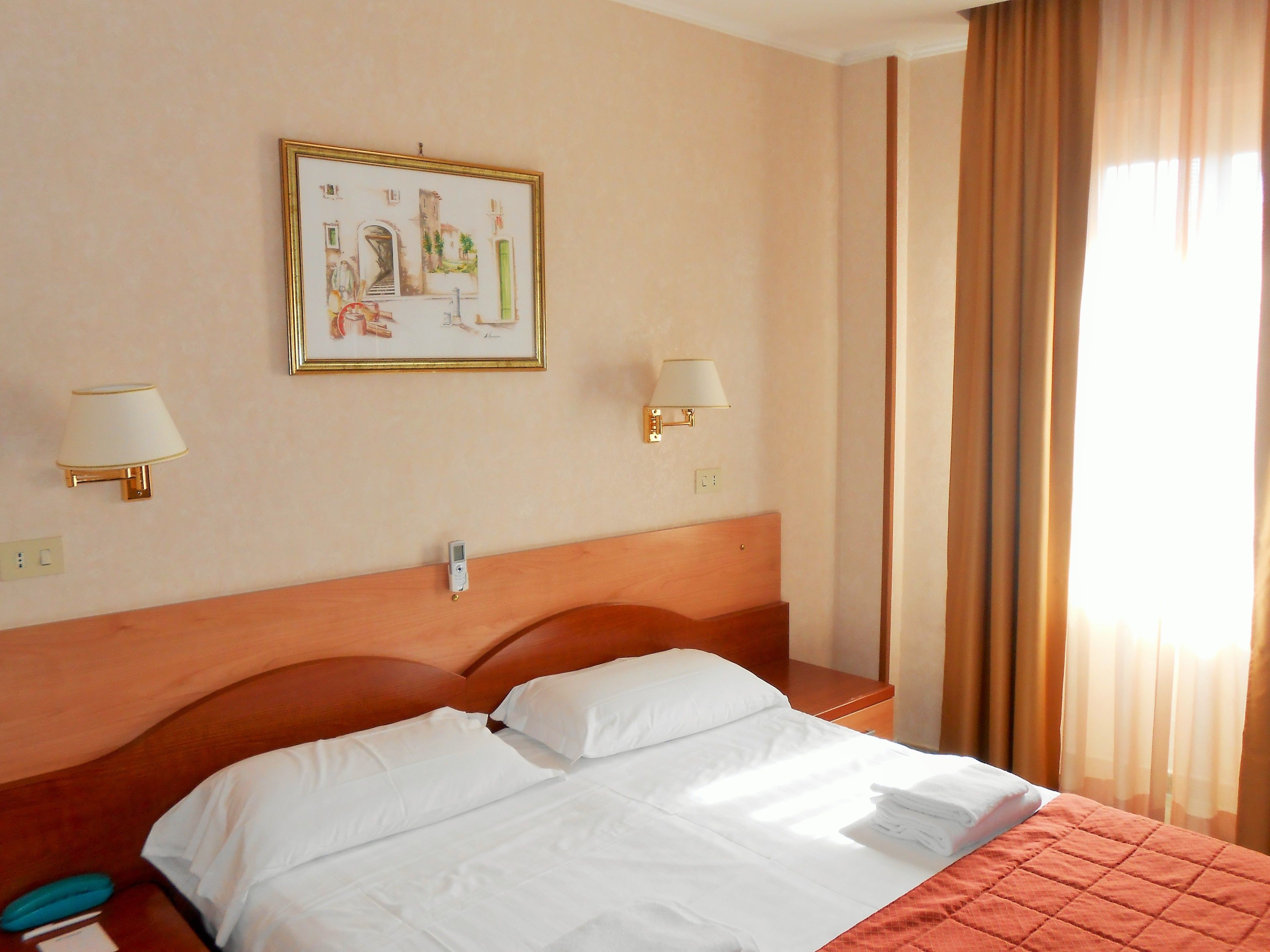 A hotel room with a large bed, two bedside lamps, a wall-mounted painting, and a window with curtains.
