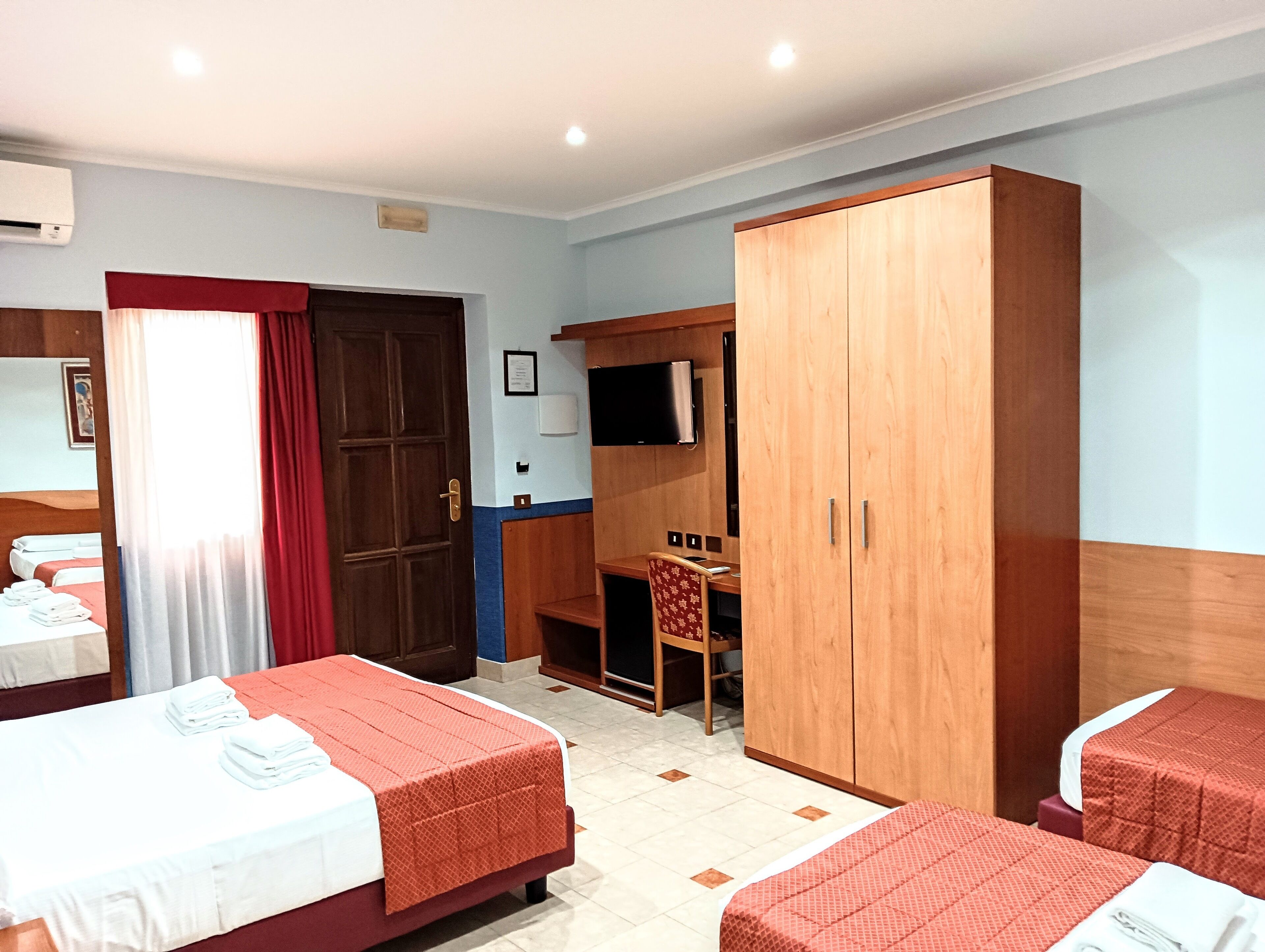 A hotel room with two beds, a wardrobe, a desk with a chair, a television, and an air conditioning unit.