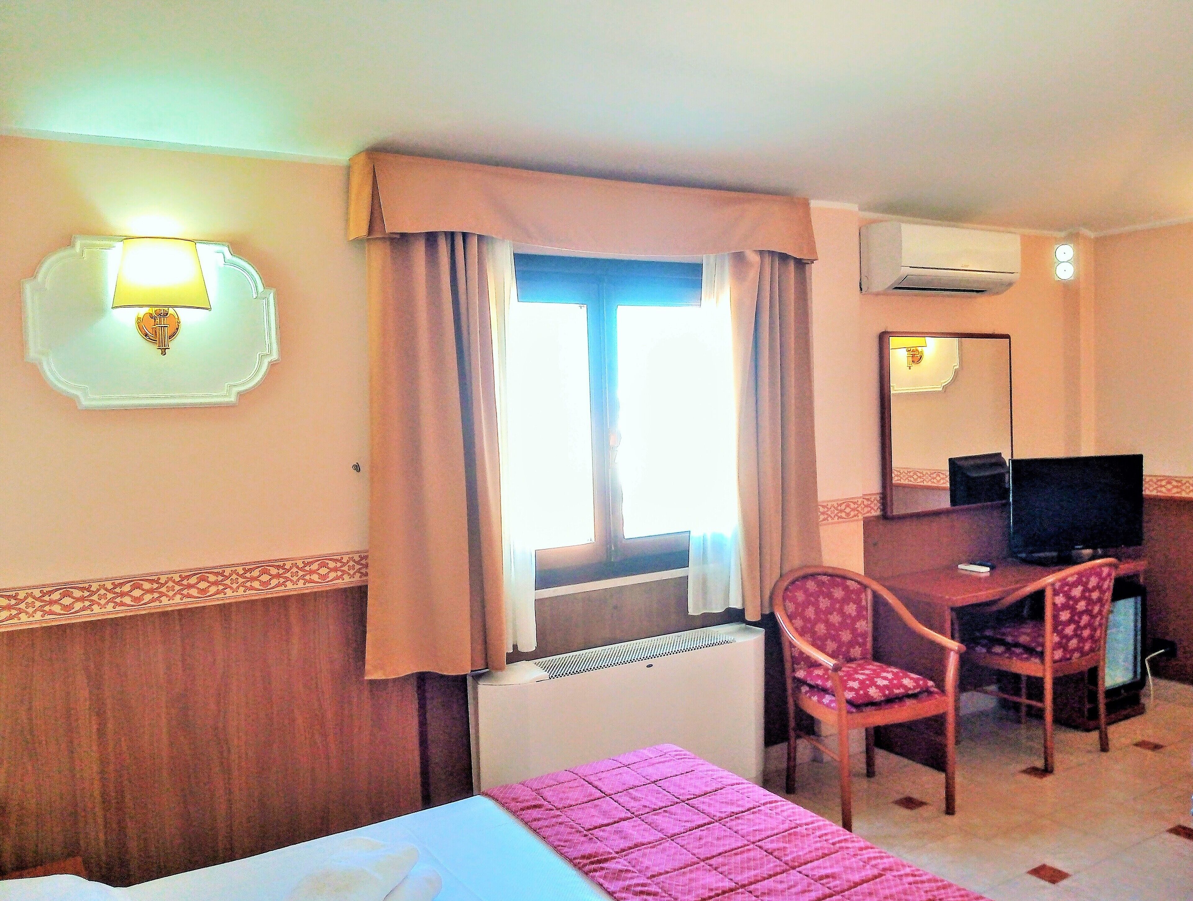 A hotel room with a bed, a desk with a chair, a television, and an air conditioning unit.